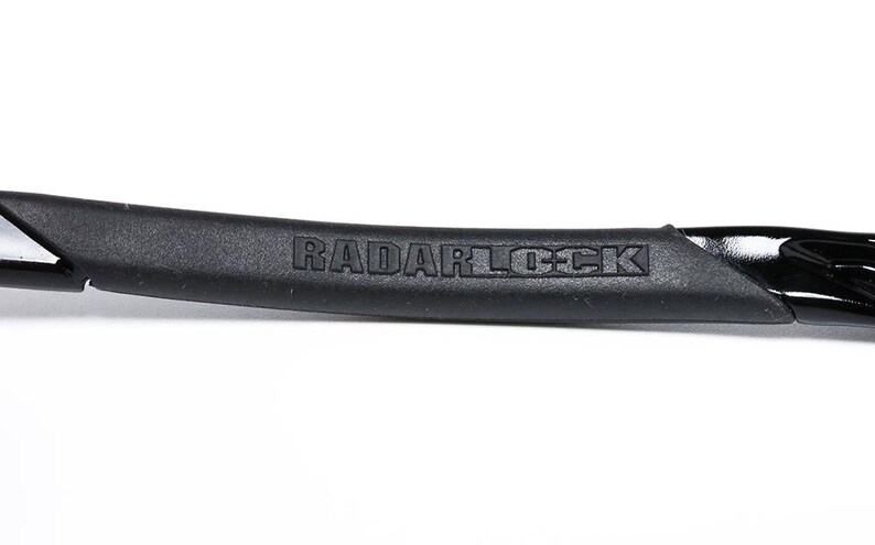 May include: Close-up of a black sunglass arm with the text "RADARLOCK" embossed on a textured rubber grip. The arm has a glossy black finish and a sleek, modern design.