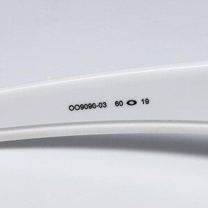 May include: Close-up of a white sunglasses arm with the numbers "OO9090-03 60 19" printed in black. The arm has a curved design and is set against a white background.
