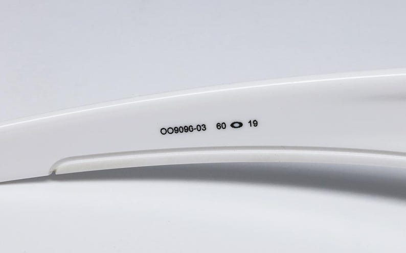 May include: Close-up of a white sunglasses arm with the numbers "OO9090-03 60 19" printed in black. The arm has a curved design and is set against a white background.