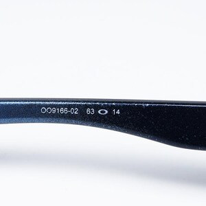 May include: Close-up of a section of a pair of blue sunglasses. The arm of the glasses displays the numbers "OO9166-02 63 O 14" in white. The word "Under" is also visible.