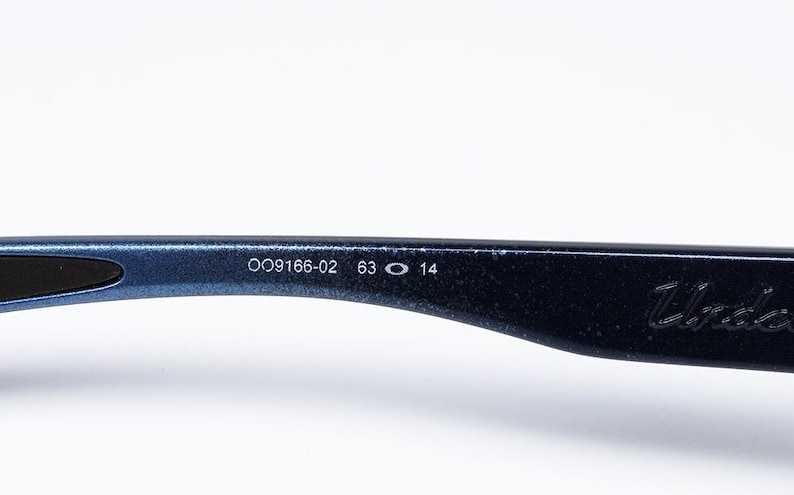 May include: Close-up of a section of a pair of blue sunglasses. The arm of the glasses displays the numbers "OO9166-02 63 O 14" in white. The word "Under" is also visible.