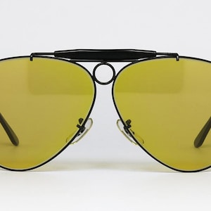 Puede incluir: Gafas de sol estilo aviador con montura negra y lentes amarillas. Las gafas tienen un doble puente y un dise&ntilde;o elegante y moderno. Las lentes son de color amarillo vibrante, ofreciendo un aspecto audaz.