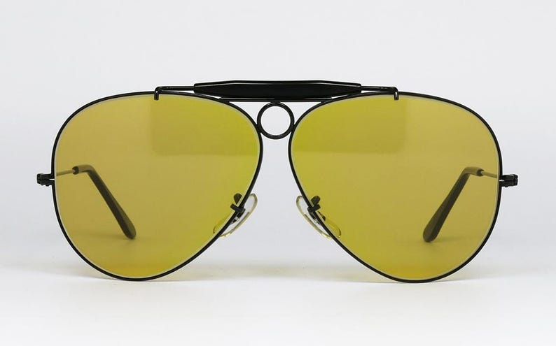 Puede incluir: Gafas de sol estilo aviador con montura negra y lentes amarillas. Las gafas tienen un doble puente y un dise&ntilde;o elegante y moderno. Las lentes son de color amarillo vibrante, ofreciendo un aspecto audaz.
