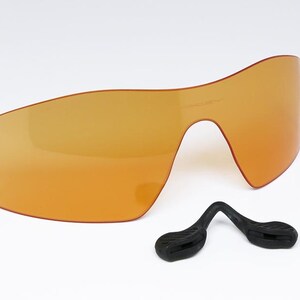 May include: An orange-tinted, wraparound style lens with a black nose piece. The lens is a single piece with a curved design, and the nose piece is a separate component with a curved shape.