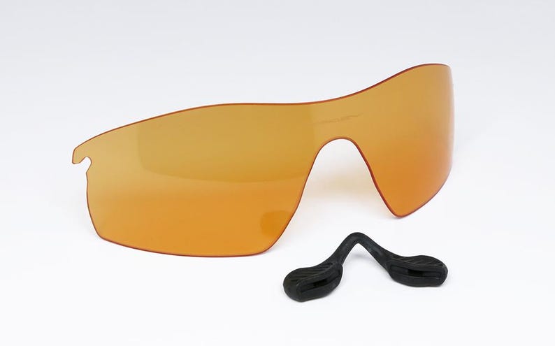 May include: An orange-tinted, wraparound style lens with a black nose piece. The lens is a single piece with a curved design, and the nose piece is a separate component with a curved shape.