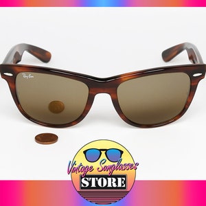 Peut inclure: Lunettes de soleil Ray-Ban Wayfarer en &eacute;caille de tortue brune avec des verres bruns. Les lunettes sont pos&eacute;es sur une surface blanche avec une pi&egrave;ce de monnaie devant elles.
