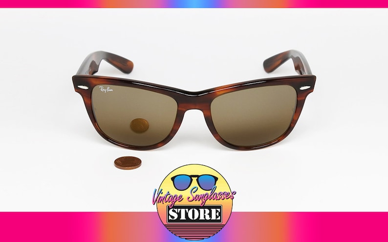 Peut inclure: Lunettes de soleil Ray-Ban Wayfarer en &eacute;caille de tortue brune avec des verres bruns. Les lunettes sont pos&eacute;es sur une surface blanche avec une pi&egrave;ce de monnaie devant elles.