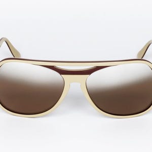 May include: Aviator-style sunglasses with a cream-colored frame and brown gradient lenses. The top bar and temples are a darker brown. The sunglasses are set against a white background.
