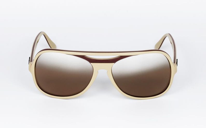 May include: Aviator-style sunglasses with a cream-colored frame and brown gradient lenses. The top bar and temples are a darker brown. The sunglasses are set against a white background.