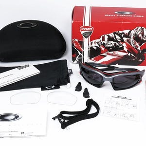Puede incluir: Un conjunto de gafas de sol Oakley Ducati Signature Series negras y rojas con lentes grises, una funda de transporte negra, una bolsa de microfibra negra, dos lentes de repuesto transparentes, una correa negra y un folleto de instrucciones negro y rojo.