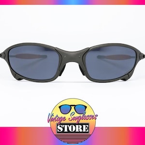 May include: Black sunglasses with dark blue lenses. The sunglasses have a matte finish and a curved design. The image includes a logo for a vintage sunglasses store.