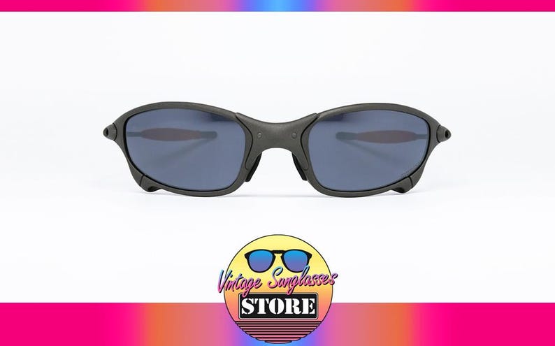 May include: Black sunglasses with dark blue lenses. The sunglasses have a matte finish and a curved design. The image includes a logo for a vintage sunglasses store.