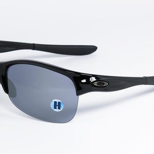 May include: Black sunglasses with gray lenses. The sunglasses have a sleek, curved design with a glossy finish. The arms curve around the ears for a secure fit. A small, blue and white sticker with the letter "H" is on the lens.