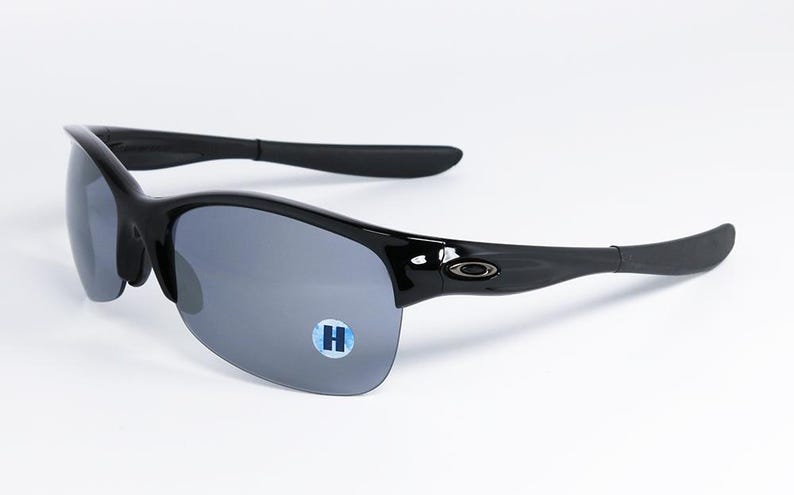 May include: Black sunglasses with gray lenses. The sunglasses have a sleek, curved design with a glossy finish. The arms curve around the ears for a secure fit. A small, blue and white sticker with the letter "H" is on the lens.