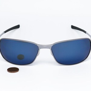 May include: Silver-framed sunglasses with blue lenses. The glasses have a rectangular shape and black arms. A penny is placed below the sunglasses for size comparison. The sunglasses are likely for outdoor use.