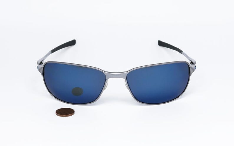 May include: Silver-framed sunglasses with blue lenses. The glasses have a rectangular shape and black arms. A penny is placed below the sunglasses for size comparison. The sunglasses are likely for outdoor use.