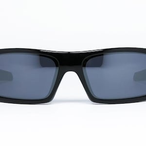 May include: Black sunglasses with a rectangular frame and gray lenses. The sunglasses have a thick, glossy frame and a modern, sporty design. The lenses are a dark gray color, providing protection from the sun.