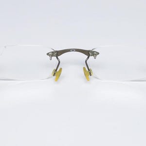 May include: A pair of rimless eyeglasses with a silver frame and clear lenses. The arms of the glasses are made of metal and have a silver finish.