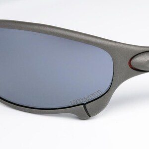 May include: Close-up of gray sunglasses with gray lenses. The sunglasses have a curved design with the word "DUCATI" printed on the frame. The frame has a matte finish and a small red accent near the temple.