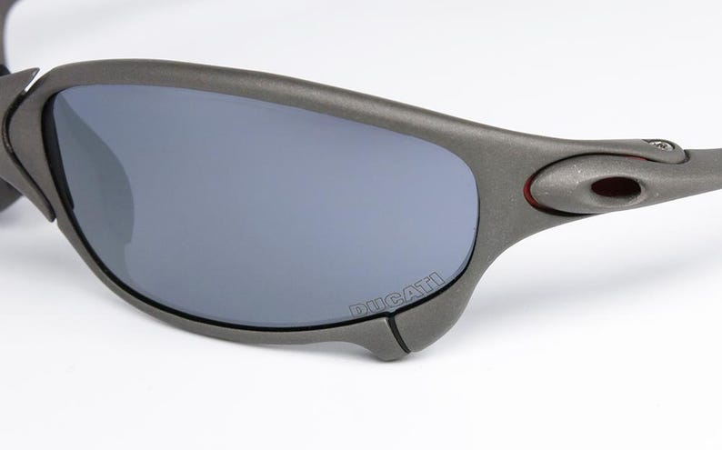 May include: Close-up of gray sunglasses with gray lenses. The sunglasses have a curved design with the word "DUCATI" printed on the frame. The frame has a matte finish and a small red accent near the temple.