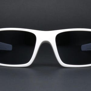 May include: White sunglasses with a thick, rectangular frame and dark lenses. The sunglasses have a modern, sporty design. The frame is a bright white colour, contrasting with the dark lenses. Suitable for outdoor activities.