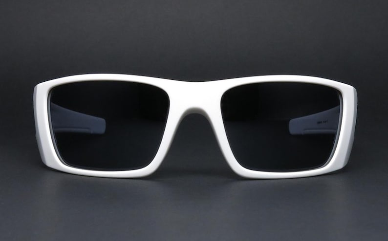 May include: White sunglasses with a thick, rectangular frame and dark lenses. The sunglasses have a modern, sporty design. The frame is a bright white colour, contrasting with the dark lenses. Suitable for outdoor activities.