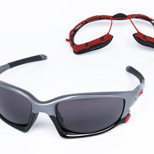 Puede incluir: Un par de gafas de sol grises con monturas negras y lentes grises oscuras. Las gafas de sol tienen detalles rojos en los lados. Tambi&eacute;n se muestra un marco rojo separado.