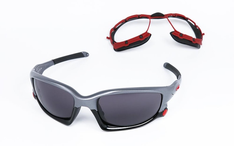 Puede incluir: Un par de gafas de sol grises con monturas negras y lentes grises oscuras. Las gafas de sol tienen detalles rojos en los lados. Tambi&eacute;n se muestra un marco rojo separado.