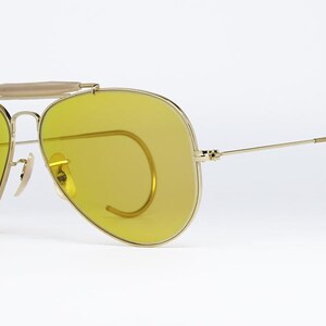 K&ouml;nnte beinhalten: Goldene Aviator-Sonnenbrille mit gelben Gl&auml;sern und einem Drahtgestell. Die Sonnenbrille hat eine markante Drahtbr&uuml;cke und einen Drahtb&uuml;gel.