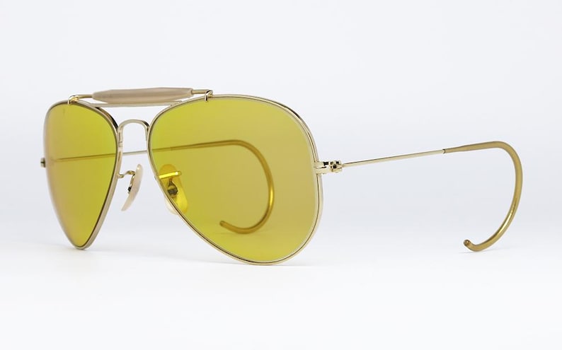 K&ouml;nnte beinhalten: Goldene Aviator-Sonnenbrille mit gelben Gl&auml;sern und einem Drahtgestell. Die Sonnenbrille hat eine markante Drahtbr&uuml;cke und einen Drahtb&uuml;gel.