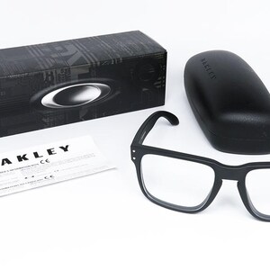 May include: Black square-framed spectacles with clear lenses, a matching black case, and a black box with the Oakley logo. A white document with the Oakley logo is also present. The spectacles are displayed on a white surface.