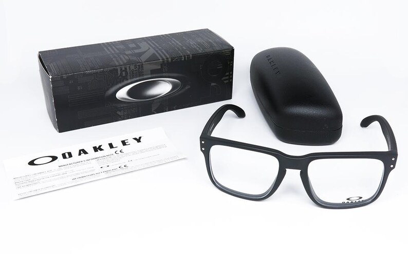 May include: Black square-framed spectacles with clear lenses, a matching black case, and a black box with the Oakley logo. A white document with the Oakley logo is also present. The spectacles are displayed on a white surface.