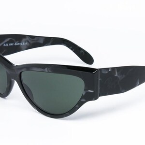 May include: Black sunglasses with a marbled pattern on the frame. The sunglasses have a cat-eye shape and green lenses. The text "BAL RAY-BAN U.S.A." is printed on the frame.