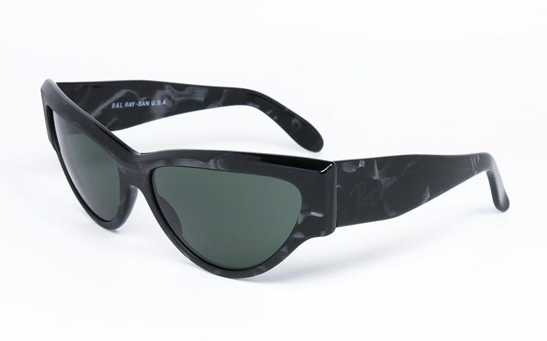 May include: Black sunglasses with a marbled pattern on the frame. The sunglasses have a cat-eye shape and green lenses. The text "BAL RAY-BAN U.S.A." is printed on the frame.