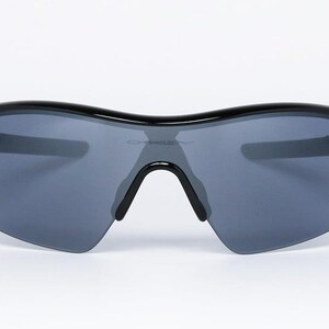 May include: Black sunglasses with gray lenses. The sunglasses have a curved top frame and a black bridge. The lenses are a dark gray color, and the frame is glossy black. The sunglasses are designed for sports and outdoor activities.