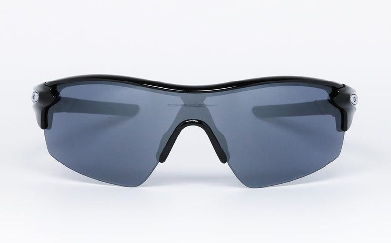 May include: Black sunglasses with gray lenses. The sunglasses have a curved top frame and a black bridge. The lenses are a dark gray color, and the frame is glossy black. The sunglasses are designed for sports and outdoor activities.
