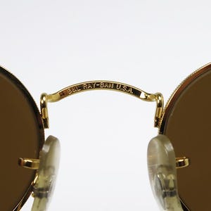 Pu&ograve; includere: Primo piano di occhiali da sole con montatura dorata e lenti marroni. Il ponte degli occhiali riporta la scritta "B&L RAY-BAN U.S.A." in nero. Le lenti sono rotonde e la montatura &egrave; un design classico.