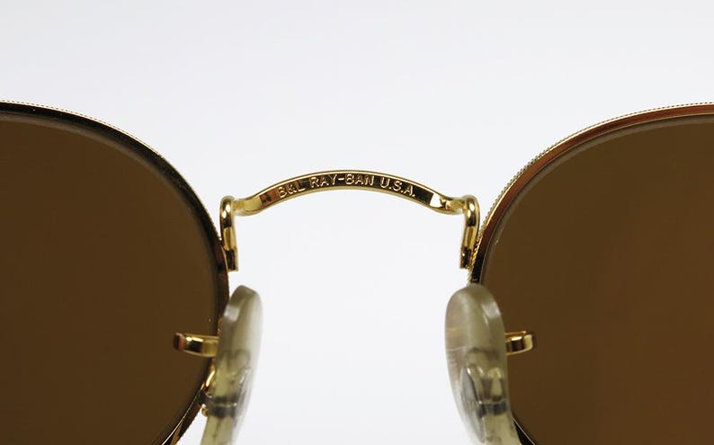 Pu&ograve; includere: Primo piano di occhiali da sole con montatura dorata e lenti marroni. Il ponte degli occhiali riporta la scritta "B&L RAY-BAN U.S.A." in nero. Le lenti sono rotonde e la montatura &egrave; un design classico.