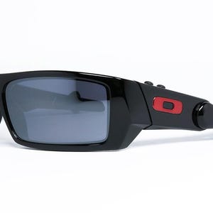 May include: Black sunglasses with gray lenses. The sunglasses have a rectangular frame with a red accent on the temple. The sunglasses are set against a white background.