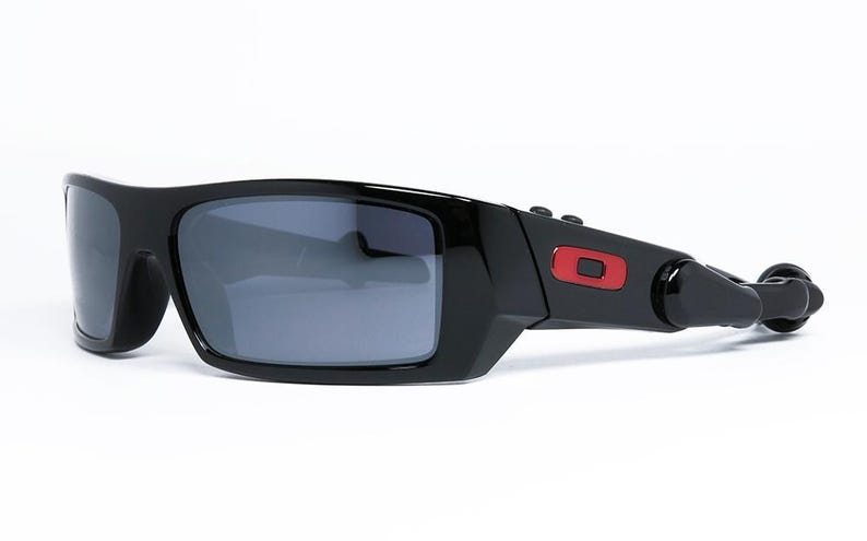May include: Black sunglasses with gray lenses. The sunglasses have a rectangular frame with a red accent on the temple. The sunglasses are set against a white background.