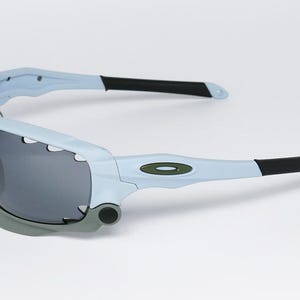 May include: Light blue and gray sports sunglasses with dark gray lenses. The sunglasses have a distinctive design with a light blue frame, gray accents, and black temple tips. The Oakley logo is visible on the temple.