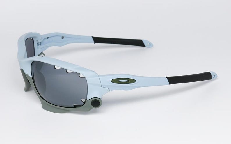 May include: Light blue and gray sports sunglasses with dark gray lenses. The sunglasses have a distinctive design with a light blue frame, gray accents, and black temple tips. The Oakley logo is visible on the temple.
