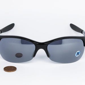 May include: Black sunglasses with gray lenses. The sunglasses have a curved frame and a small blue sticker with a white "H" on the right lens. A penny is next to the sunglasses.
