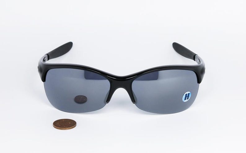 May include: Black sunglasses with gray lenses. The sunglasses have a curved frame and a small blue sticker with a white "H" on the right lens. A penny is next to the sunglasses.