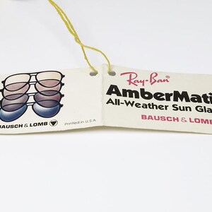 Puede incluir: Una etiqueta vintage de gafas de sol Ray-Ban AmberMatic All-Weather. La etiqueta presenta un gr&aacute;fico de gafas de sol en capas y el logotipo de Ray-Ban. Tambi&eacute;n se ve el texto "AmberMatic" y "Bausch & Lomb".