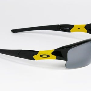 K&ouml;nnte beinhalten: Schwarze Sonnenbrille mit grauen Gl&auml;sern und gelben Akzenten. Die Sonnenbrille hat ein schlankes, modernes Design mit einem schwarzen Rahmen und gelben Details an den B&uuml;geln. Die Gl&auml;ser sind dunkelgrau.
