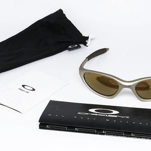 May include: Silver-framed sunglasses with brown lenses, a black drawstring pouch, and a booklet. The sunglasses have a sleek, modern design. The booklet and pouch feature the Oakley logo.