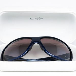 May include: Navy blue sunglasses with gradient lenses rest inside a white case. The case is open, revealing the sunglasses' dark frames and lenses. The Oakley logo is visible on the case. The sunglasses are a sporty style.