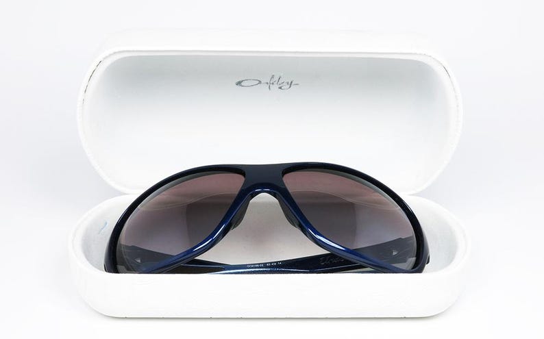 May include: Navy blue sunglasses with gradient lenses rest inside a white case. The case is open, revealing the sunglasses' dark frames and lenses. The Oakley logo is visible on the case. The sunglasses are a sporty style.