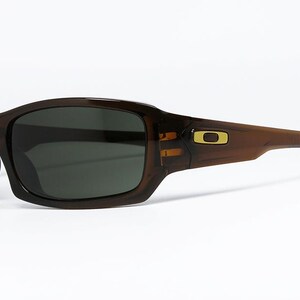 May include: Brown sunglasses with dark lenses. The sunglasses have a rectangular shape with a curved frame. A gold-coloured logo is visible on the temple. The sunglasses are designed for outdoor use.